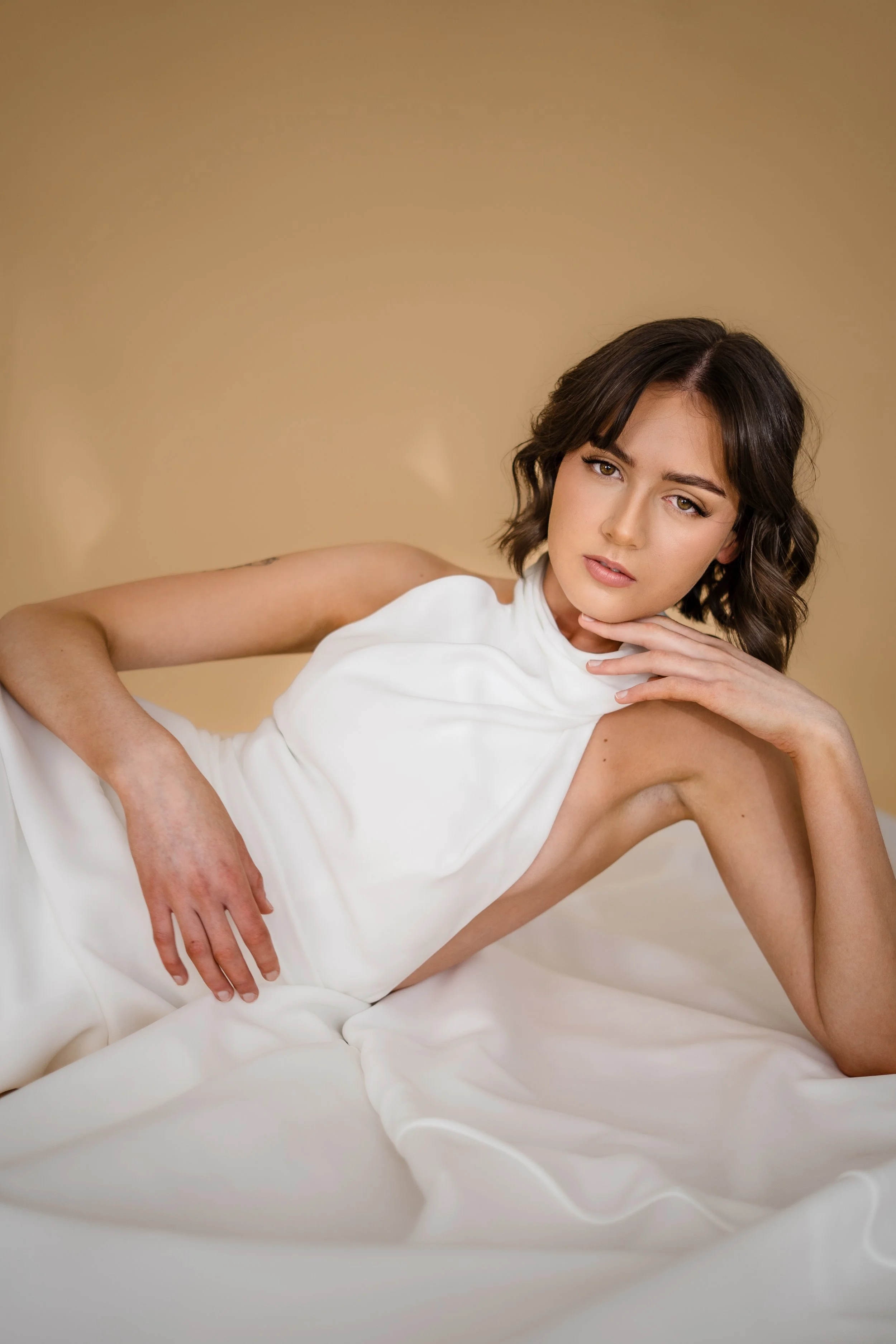 A woman with dark hair styled in loose waves, wearing a white garment, poses with her head resting on her hand against a neutral beige background. She gazes directly into the camera with a subtle expression.
