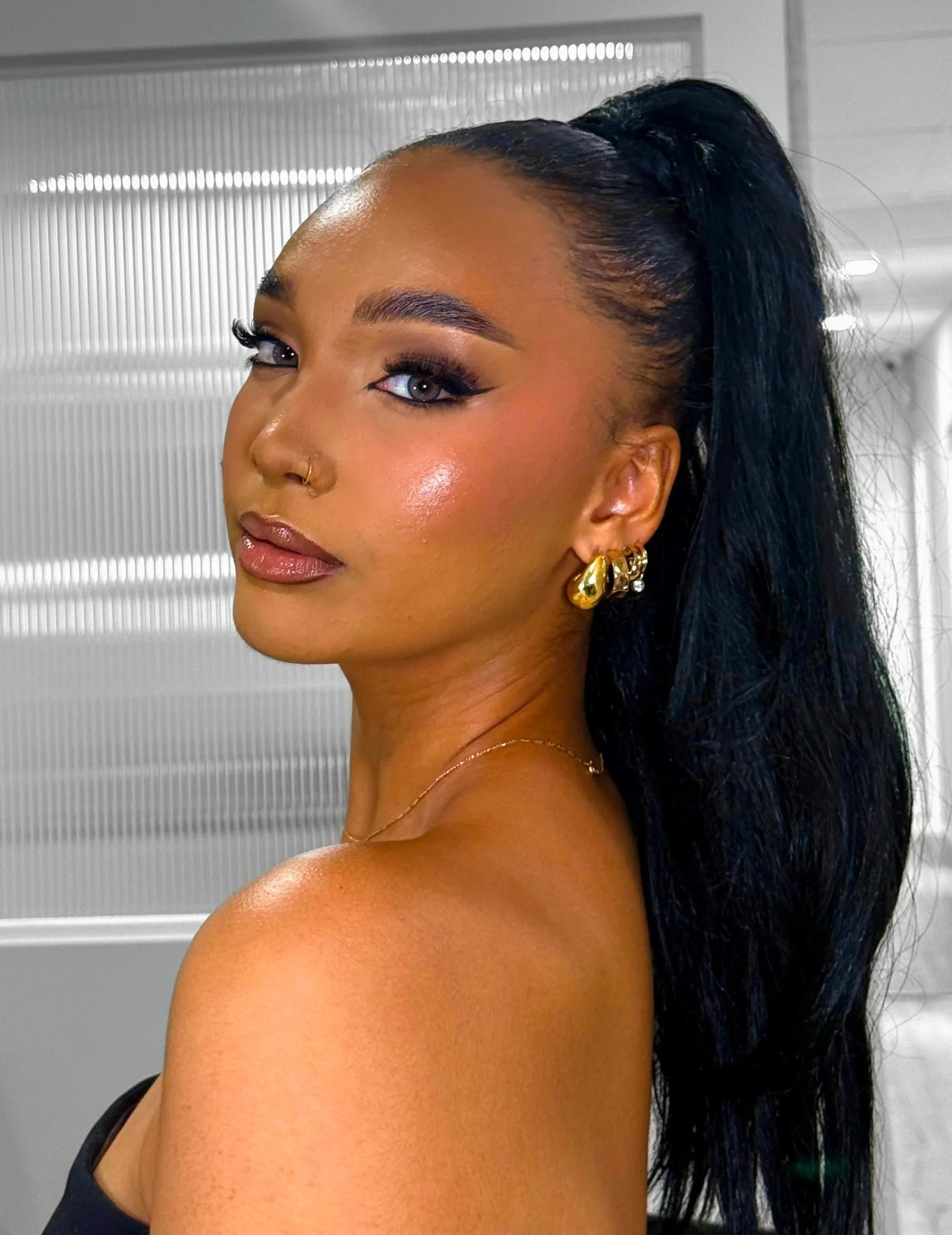 Close-up of a woman with dark hair styled in a high ponytail, wearing gold jewelry, and makeup, standing indoors in front of a metallic background.
