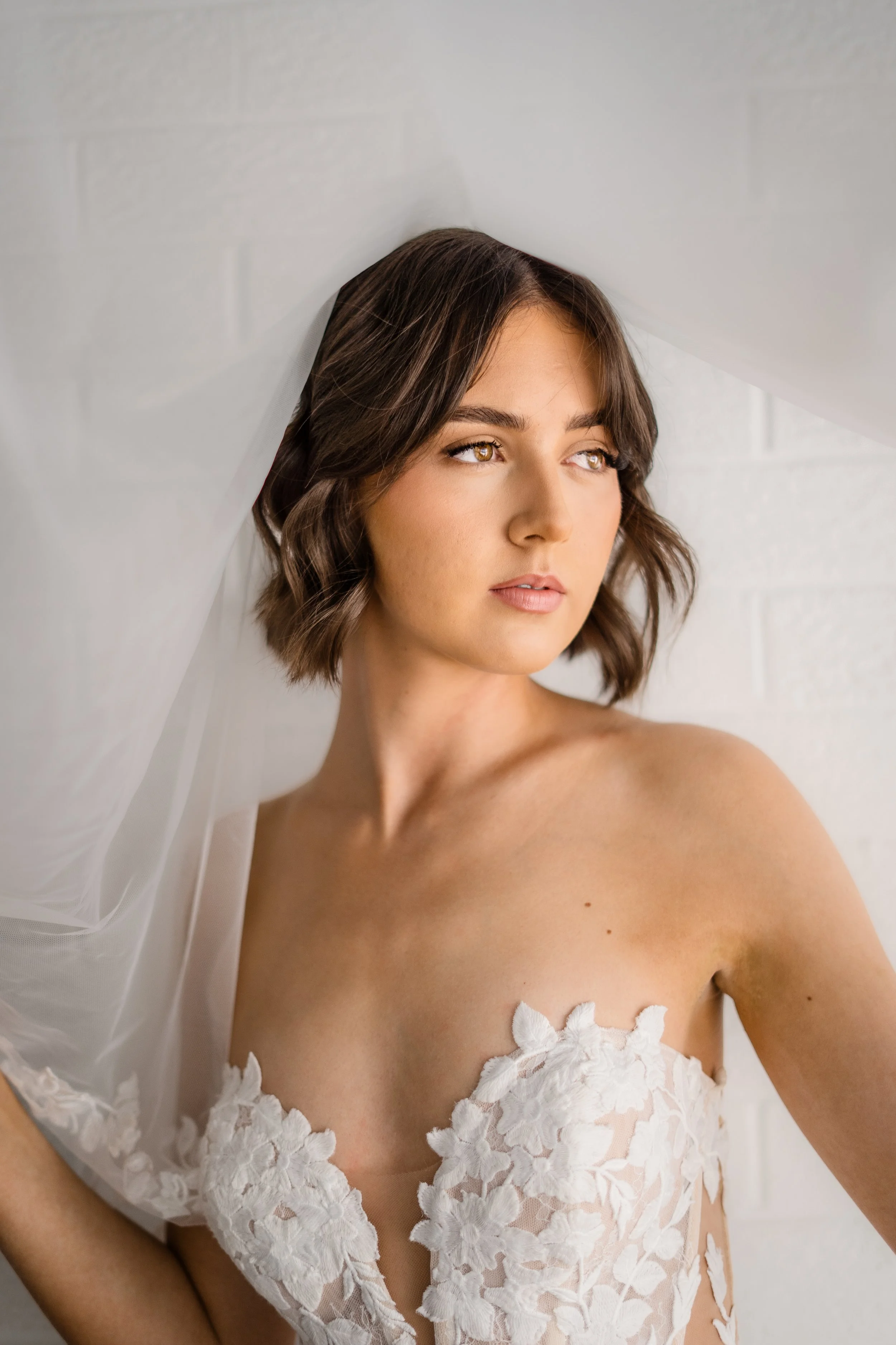 A woman with short brown hair and hazel eyes wearing a white lace dress with a floral pattern, looking off to the side against a white brick wall.