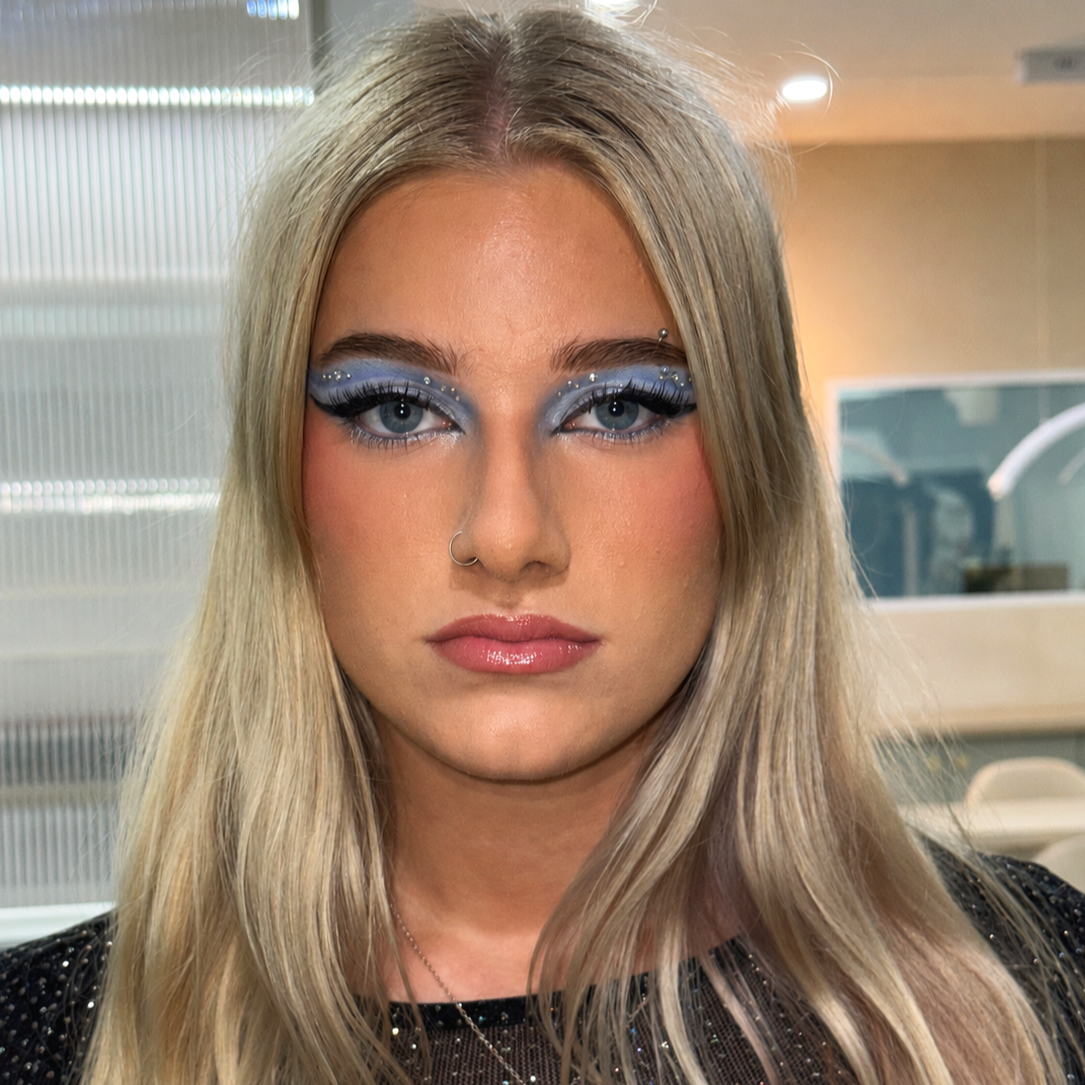 A woman with long blonde hair, blue eyes, and elaborate blue eyeshadow with rhinestones, wearing a black top and a nose piercing, indoors with blurred background.