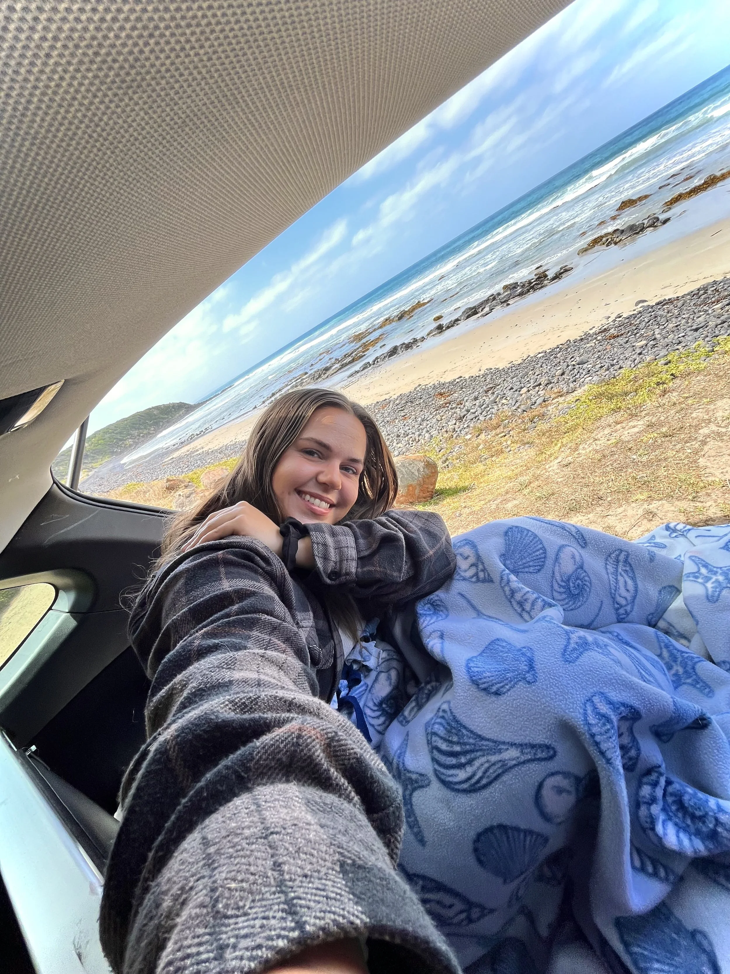 A smiling woman with brown hair sitting in the back of a vehicle, looking out the open door at a beach with waves, rocks, and a partly cloudy blue sky.