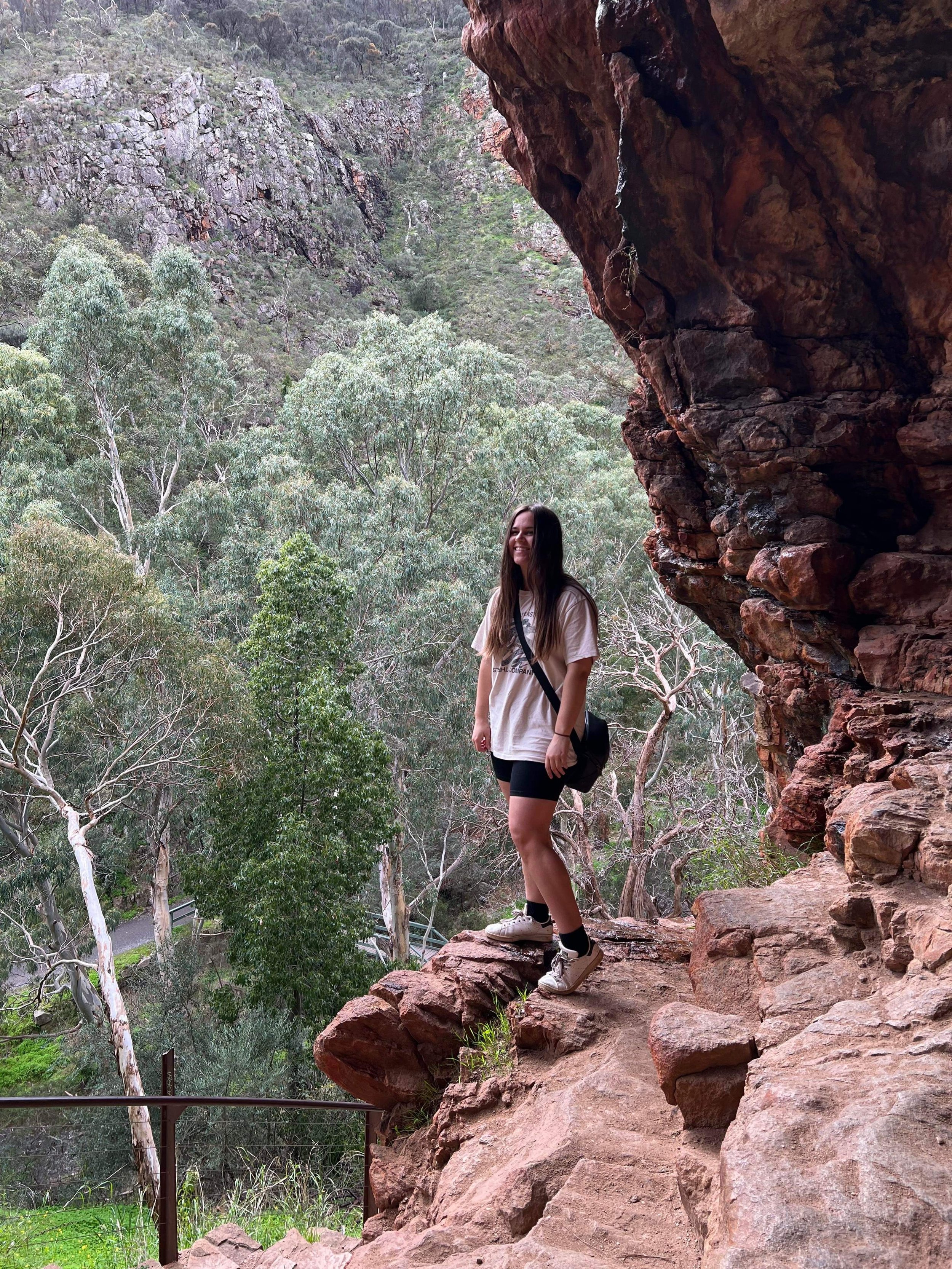 Young woman standing on rocky ledge overlooking a forested canyon with cliffs and trees in the background.