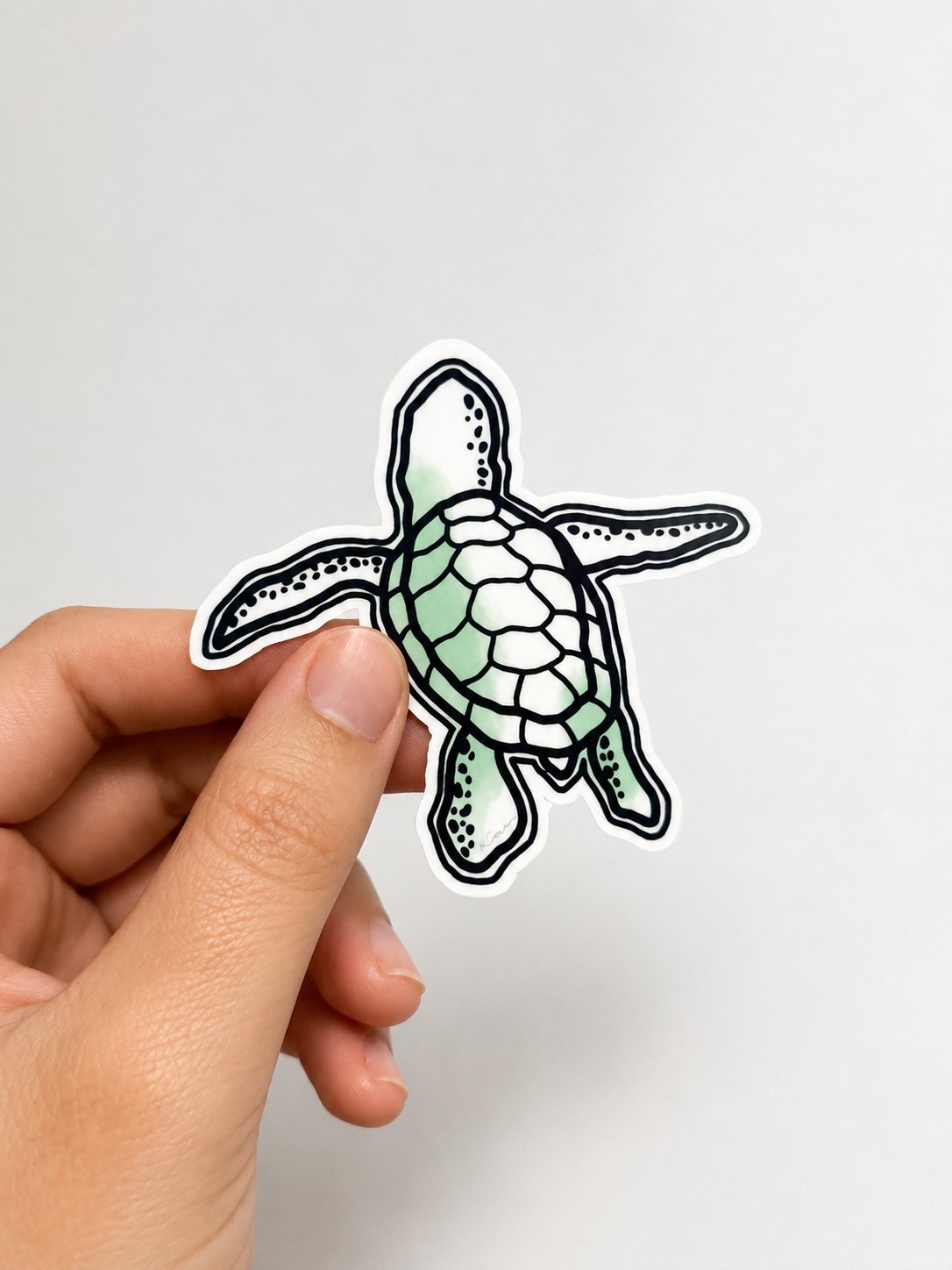 A hand holding a cutout sticker of a cartoon-style sea turtle with black outlines and some green coloring on its shell and limbs against a plain white background.