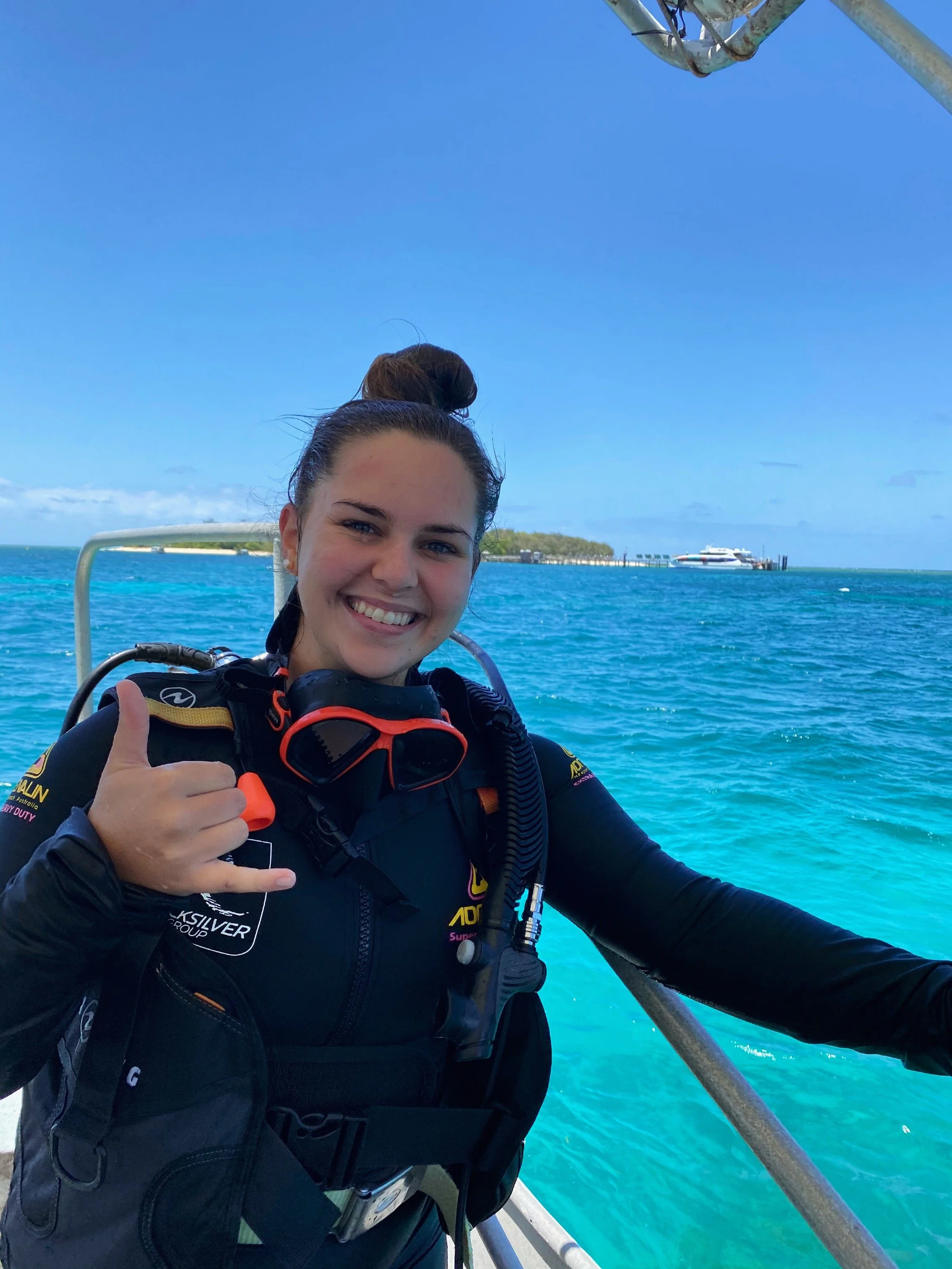 A smiling woman in a black wetsuit giving a thumbs-up while on a boat in turquoise ocean water, with a distant landmass and clear blue sky in the background.
