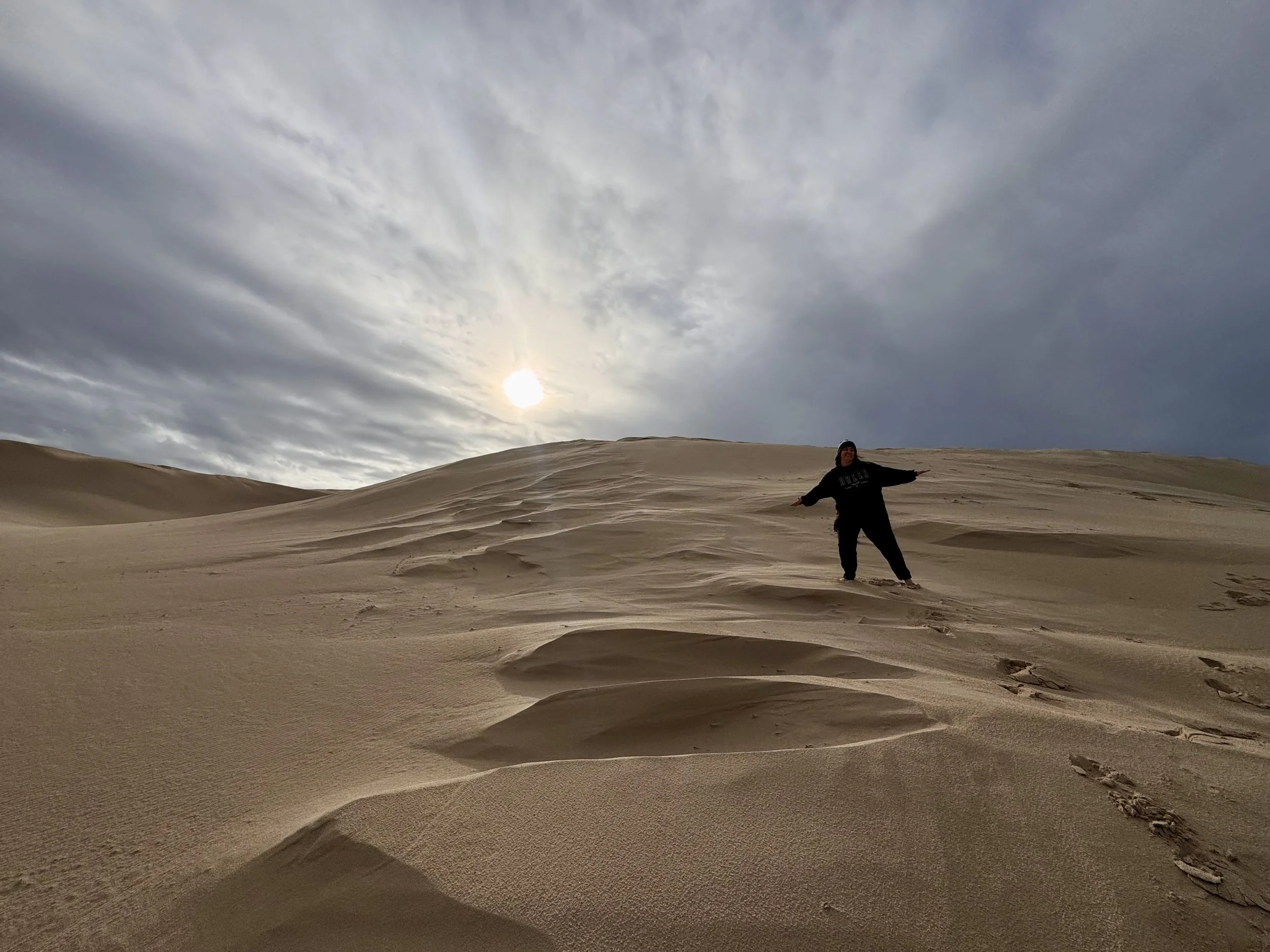A person dressed in black standing in a desert with rolling sand dunes and a cloudy sky, with the sun partially visible.