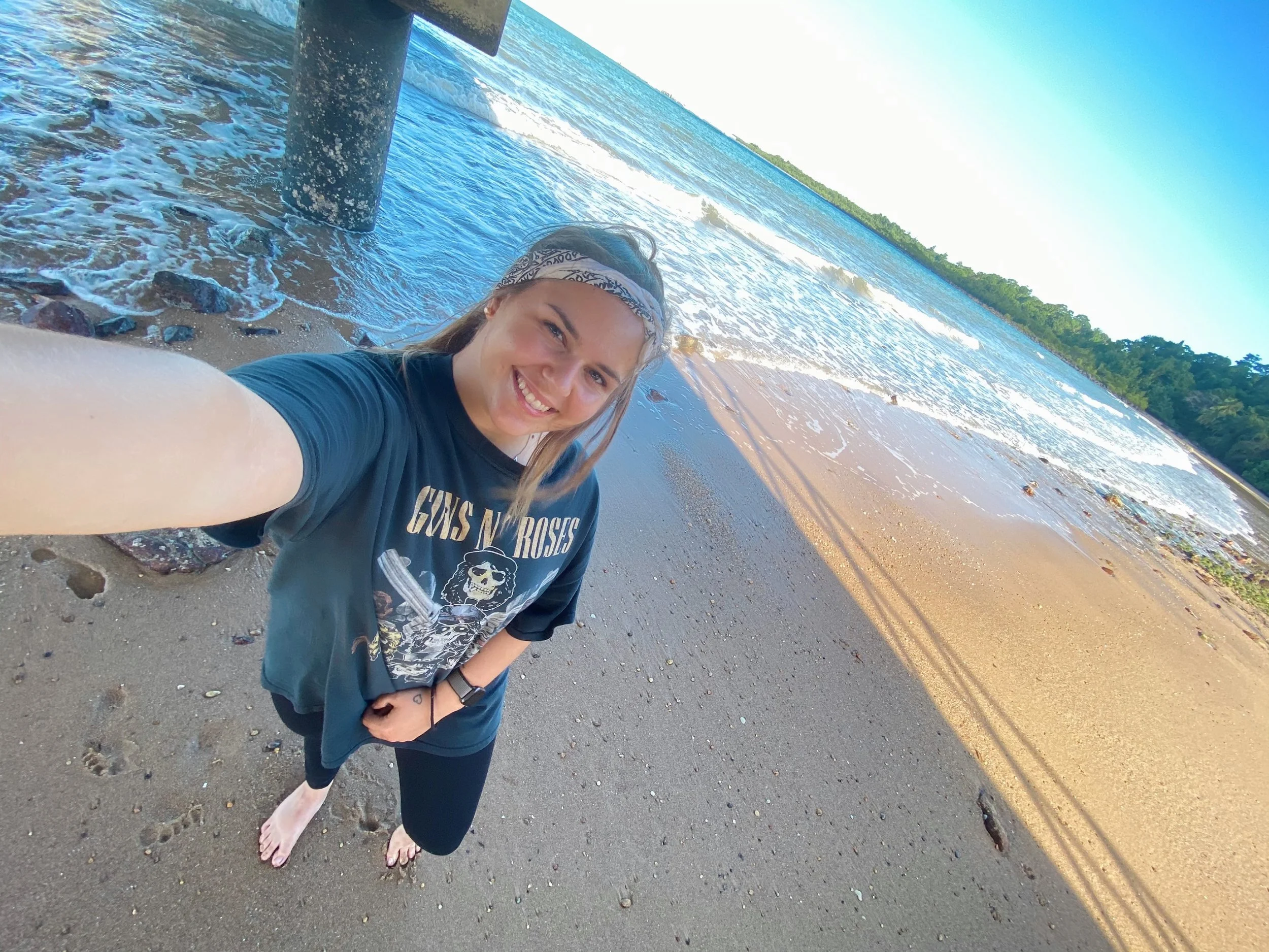 A woman with long hair, wearing a bandana, taking a selfie on a beach with waves and greenery in the background, barefoot on the sand.