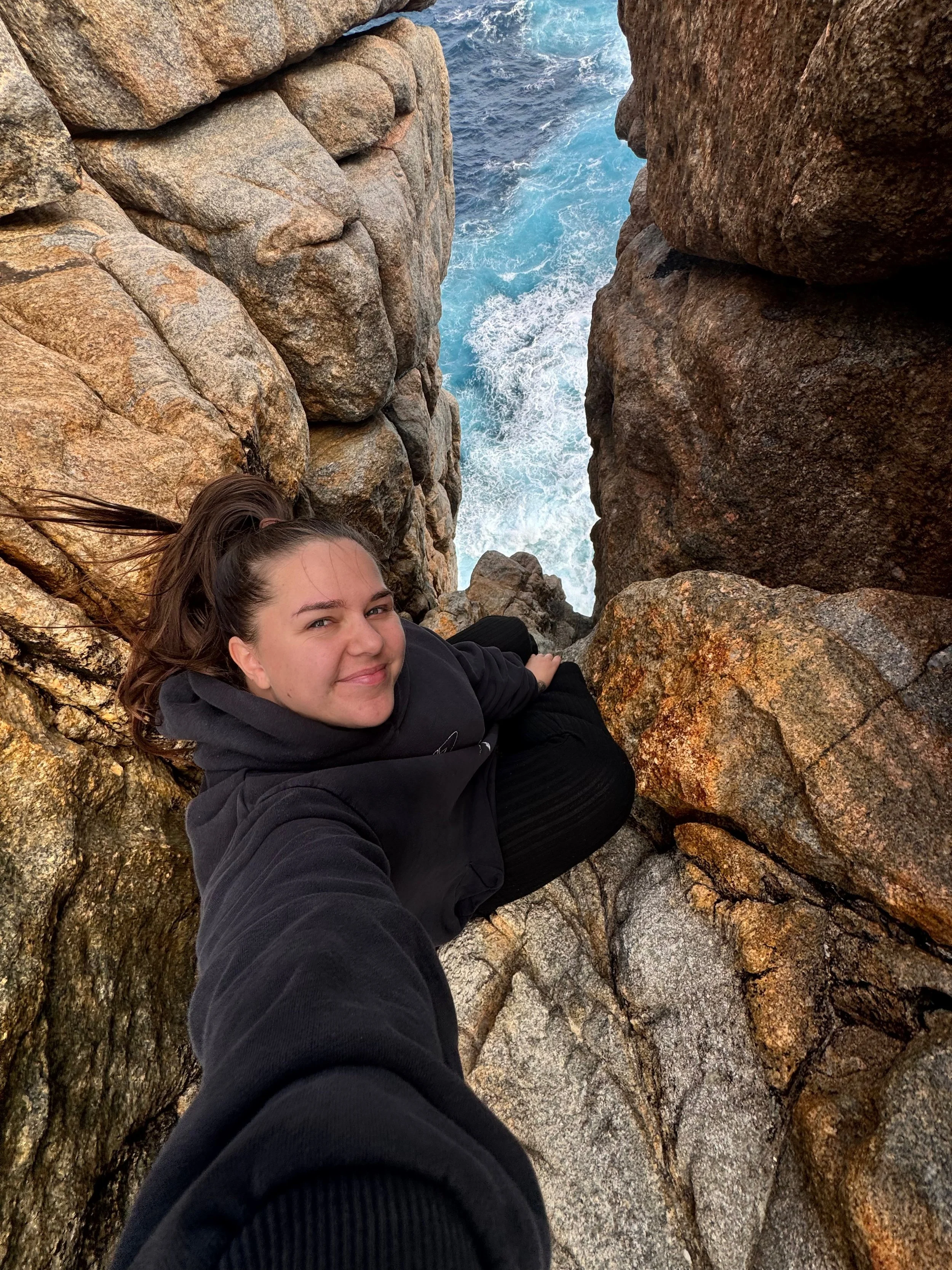 A woman with brown hair in a ponytail smiling while sitting among large rocks at the edge of a body of water, with waves visible.