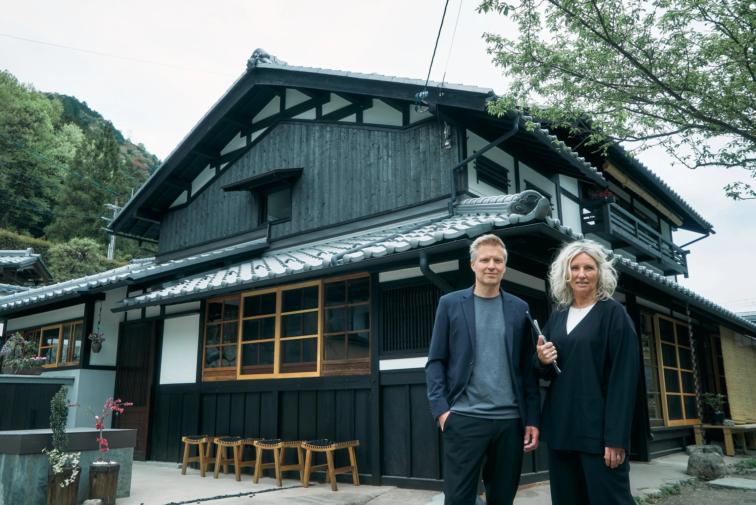 A man and woman standing outside a traditional fully renovated Japanese style kominka with a kawara tiled roof, wooden windows, and a black and white exterior, surrounded by mountains, greenery and trees.