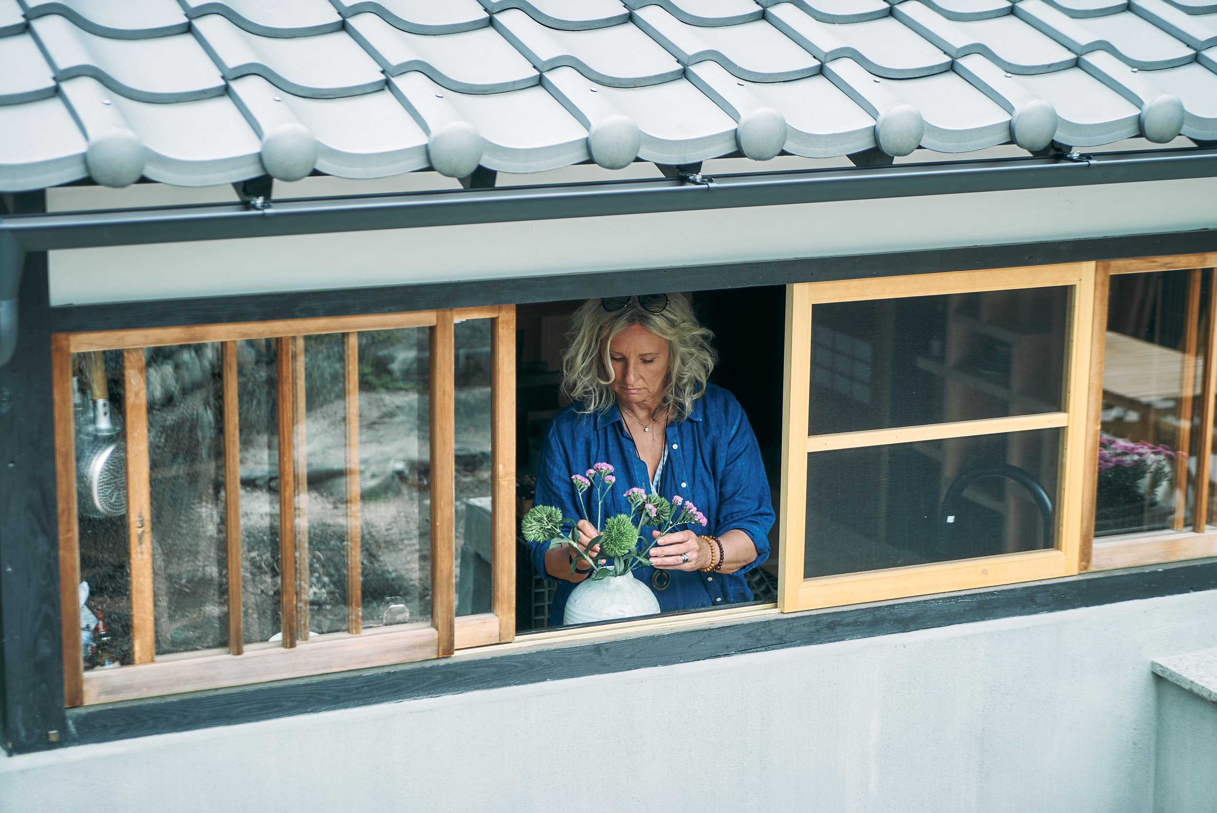 A woman with blonde hair, wearing an indigo natural linnen blue shirt, arranging flowers inspired by ikebana inside a white vase on a window ledge.