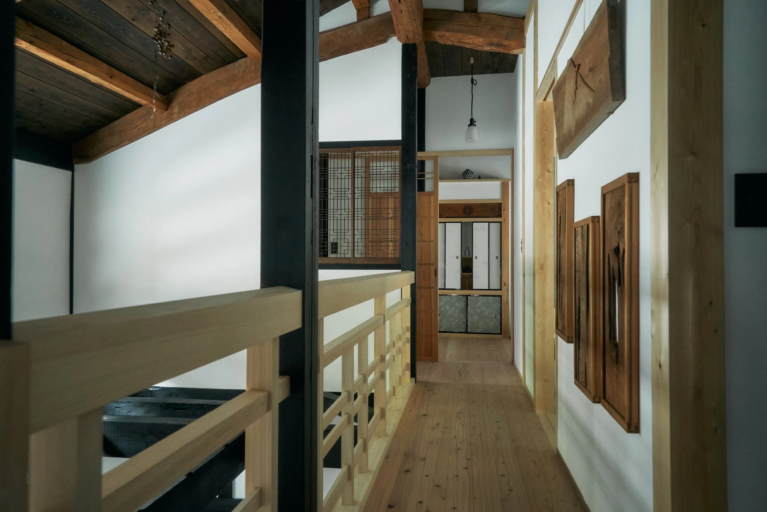 Interior of a house with wooden beams, bamboo doors, and a staircase with wooden railing. The ceiling has dark yakisugi panels, and the walls are white shikkui. A hanging vintage light bulb is visible, and there is a butsudan in the background.