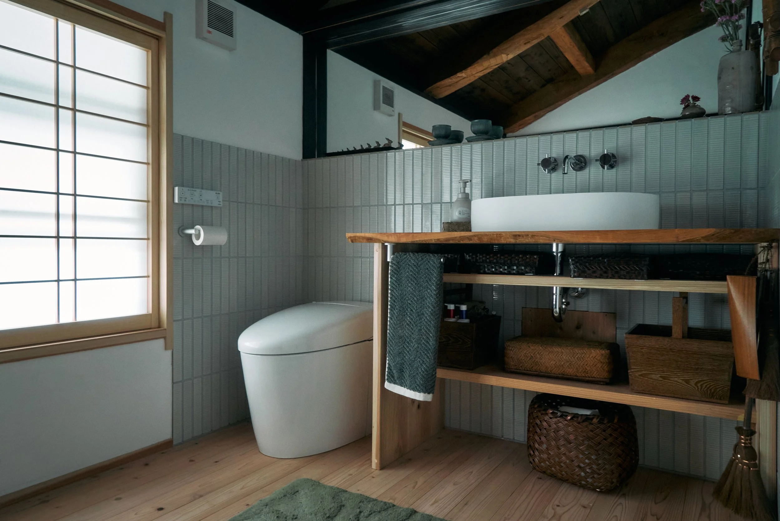 A modern bathroom with natural wood accents, a white washlet, a wooden shelf with a soap dispenser, a towel, and storage baskets, illuminated by natural light from a large window with shoji.