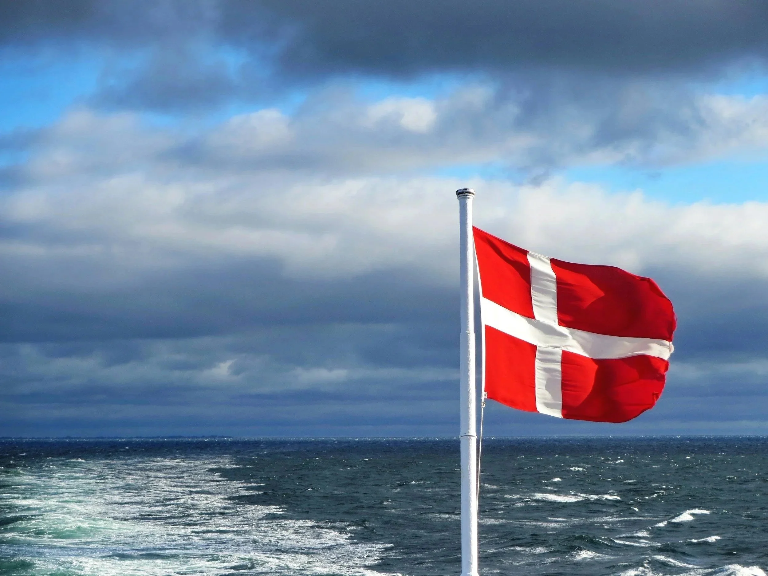A red and white striped flag mounted on a white pole flying over a body of water with waves, under a cloudy sky.