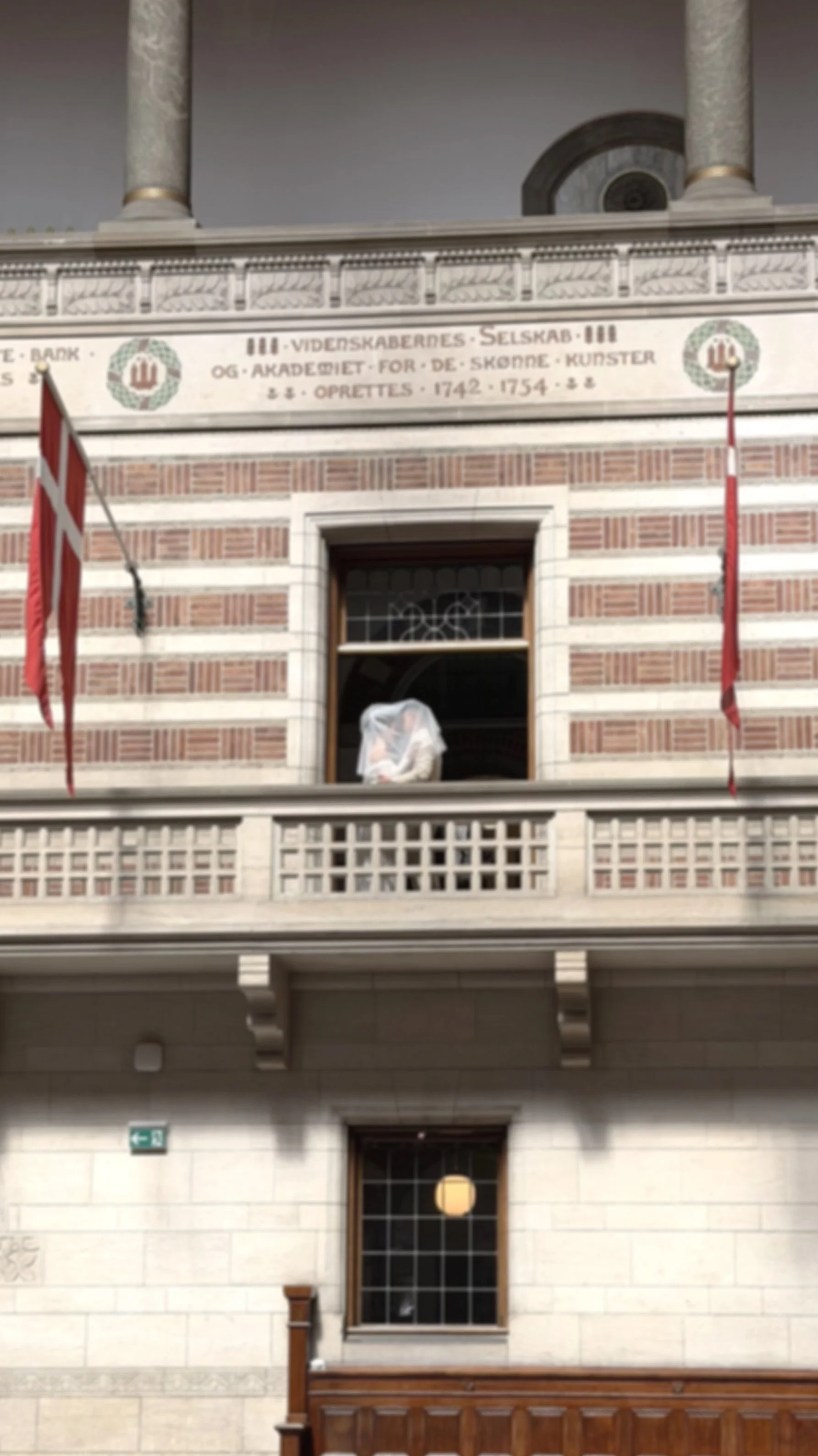 Three floors of a building with traditional architecture, featuring brick and stonework. The upper floor has a stone balcony and two flags, one Danish and one possibly Norwegian. The middle floor has a window with a protective covering, and the lower floor has a window with lanterns and a decorative stone plaque.