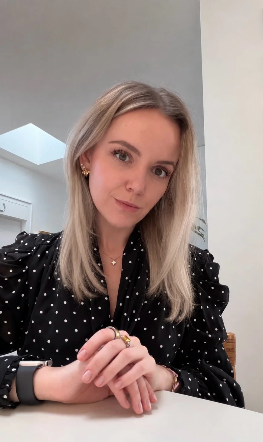 A woman with blonde hair, wearing a black polka-dot blouse, jewelry, and a smartwatch, sitting at a table in an indoor space with modern decor.