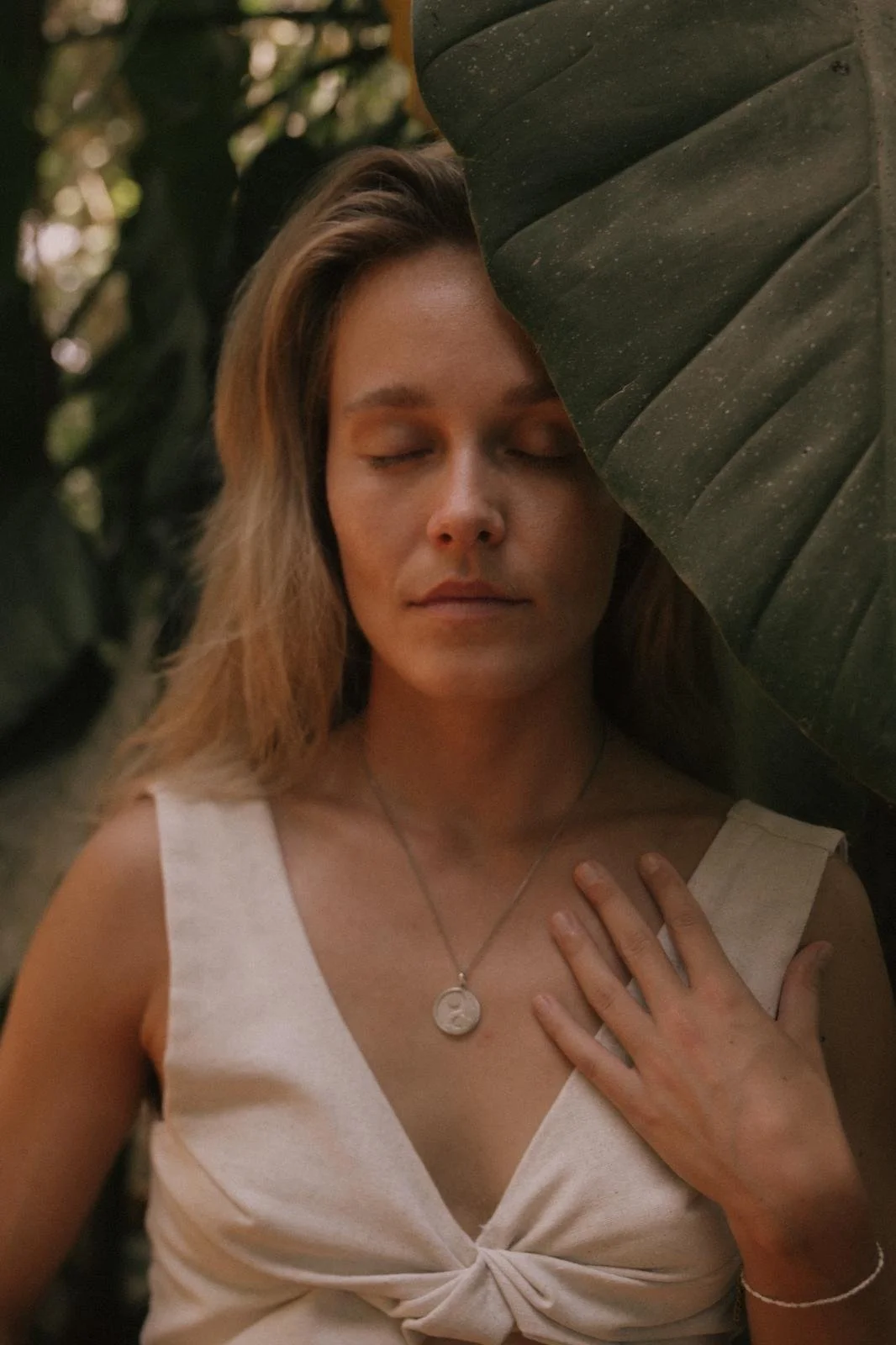 Lara (Breathwork Teacher) mit geschlossenen Augen, die einen großen grünen Blattrand teilweise vor ihrem Gesicht hat, in einem natürlichen, grünen Umfeld.