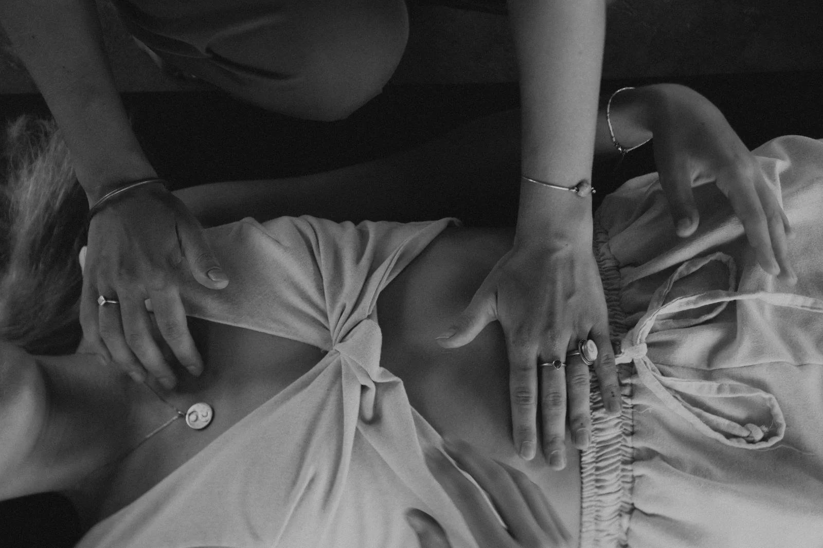 Schwarz-weiß-Foto von zwei Personen, eine von ihnen liegt auf einer Yoga Matte, die andere sitzt neben ihr und hat ihre Hände leicht über ihr schwebend. Beide tragen Schmuck und lockere Kleidung.