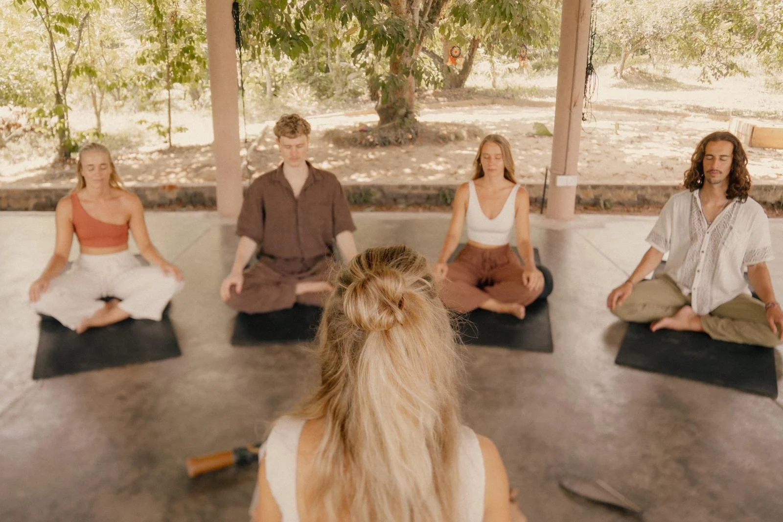 Menschen sitzen im Lotossitz mit geschlossenen Augen in einer Breathwork-Sitzung im Freien unter einem Dach mit Bäumen im Hintergrund, Lara sitzt vor ihnen zur Anleitung.