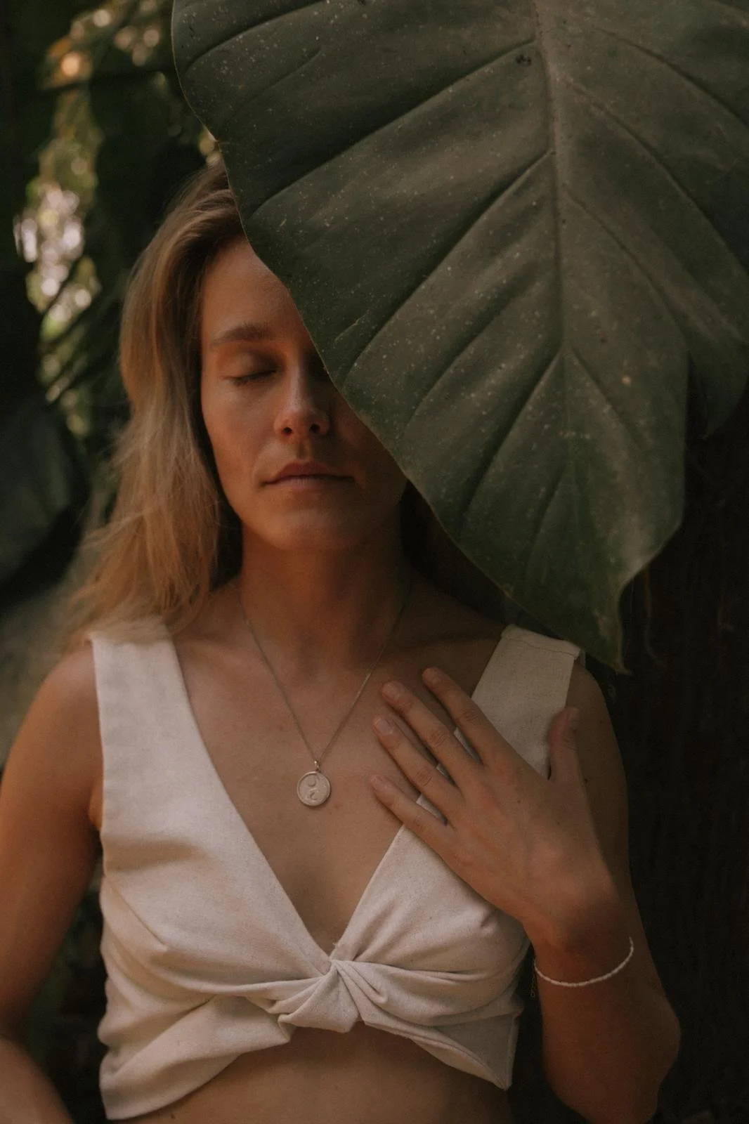 Lara (Breathwork Teacher) mit geschlossenen Augen, die einen großen grünen Blattrand teilweise vor ihrem Gesicht hat, in einem natürlichen, grünen Umfeld.