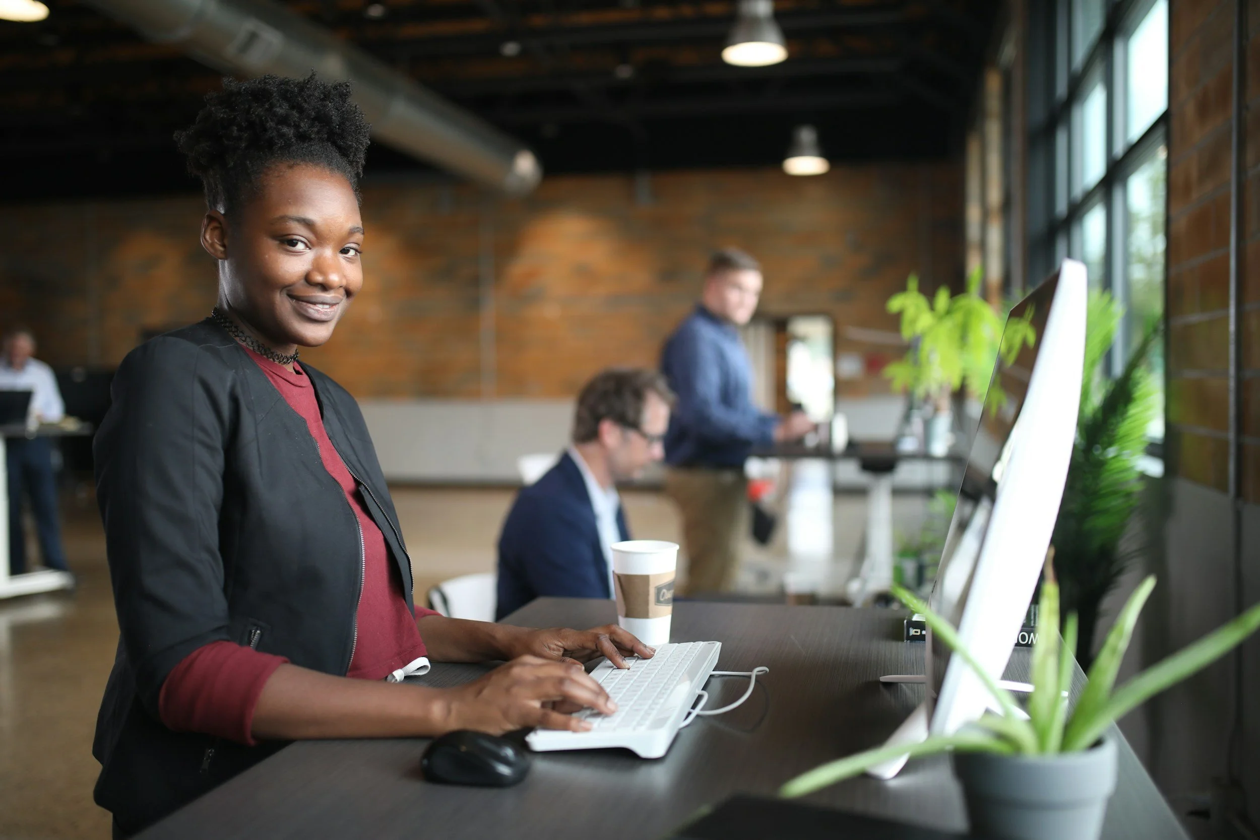 A woman with dark skin, curly hair in a bun, wearing a black jacket and red shirt, smiling while working on a white keyboard at a desk in a modern office space.