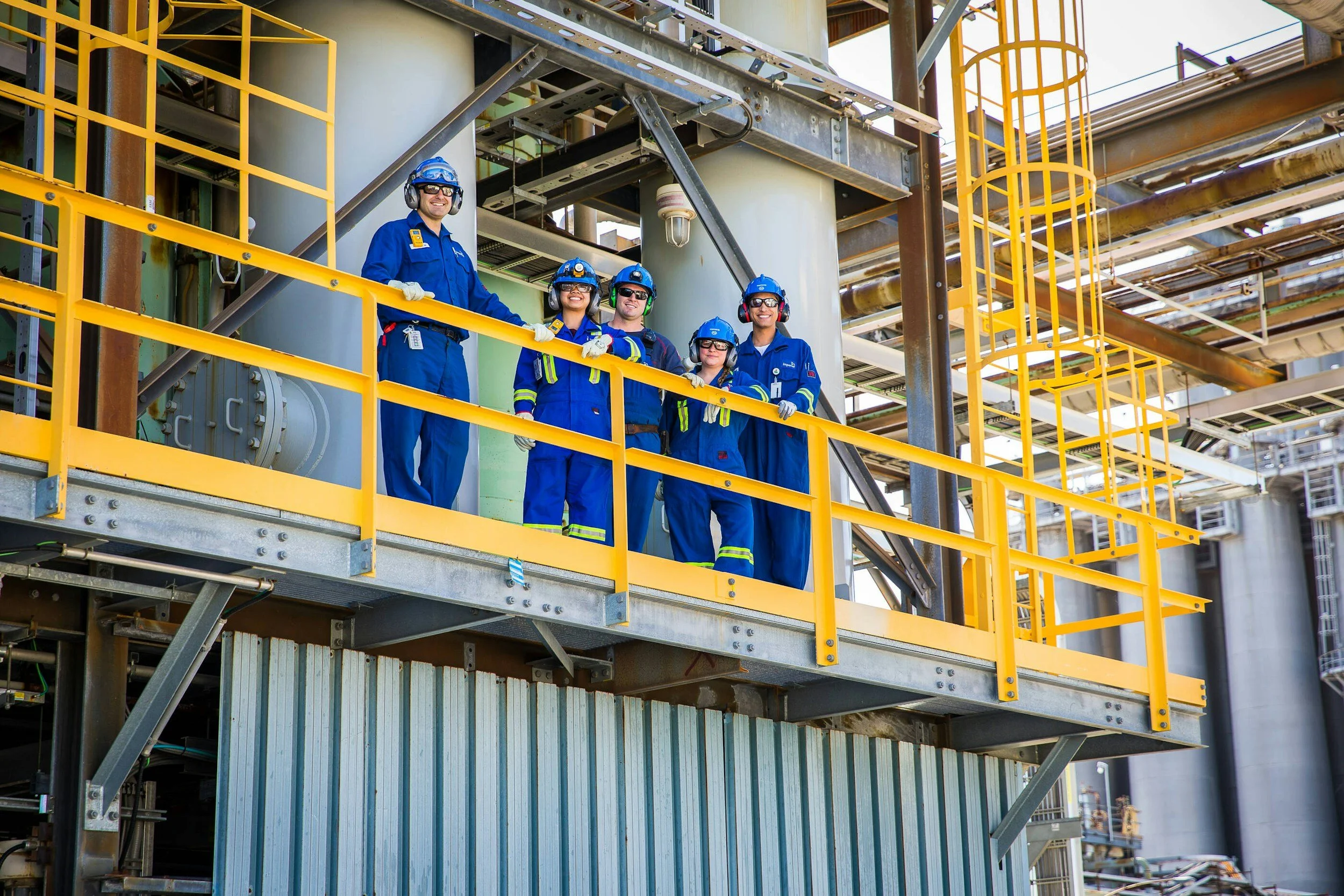 Five industrial workers wearing blue coveralls, safety helmets, and ear protection standing on a yellow safety railing at an industrial plant.