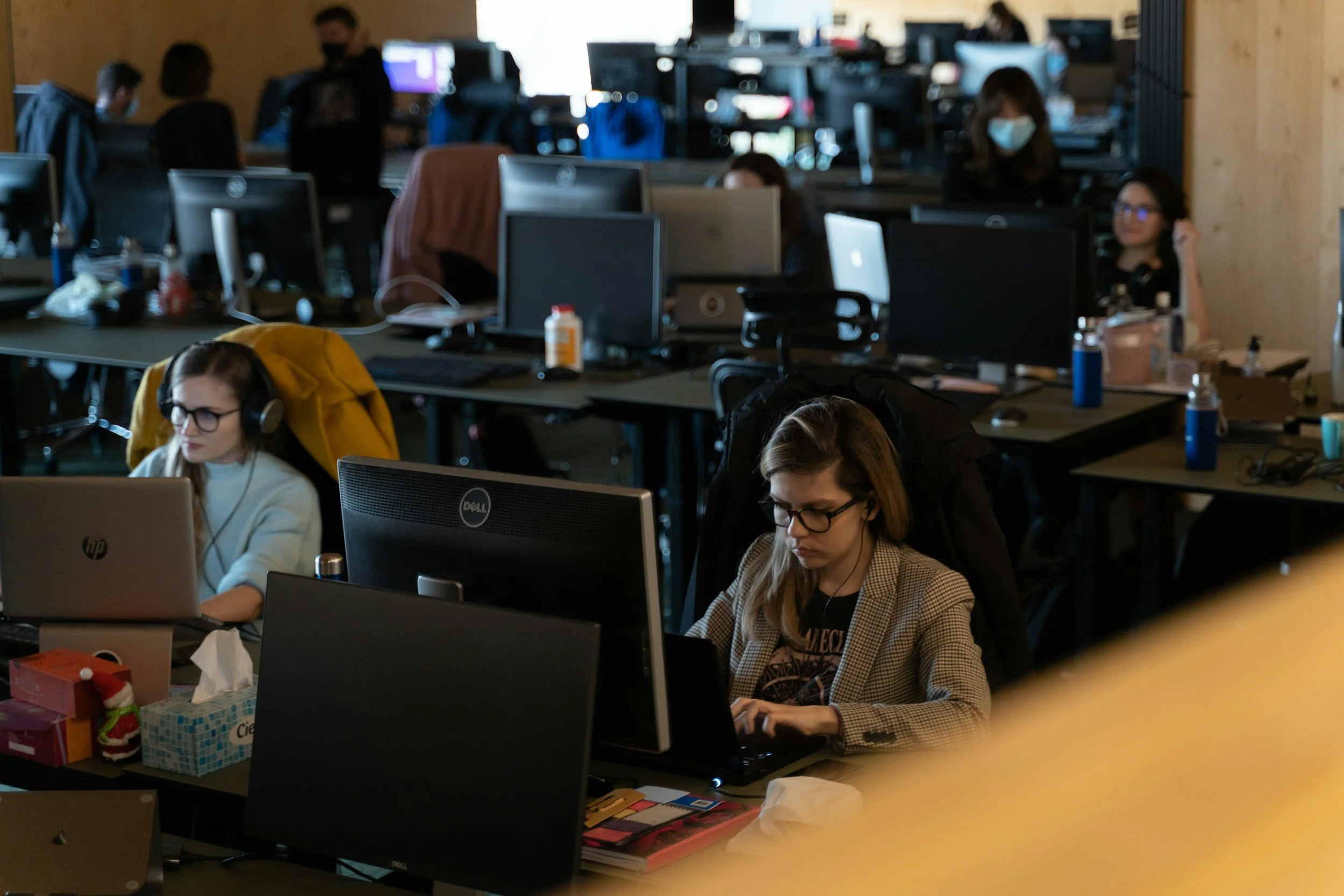 A busy office with multiple people working at desks with computers. Some people are wearing masks, and the workspace has clutter like tissue boxes, bottles, and snacks.