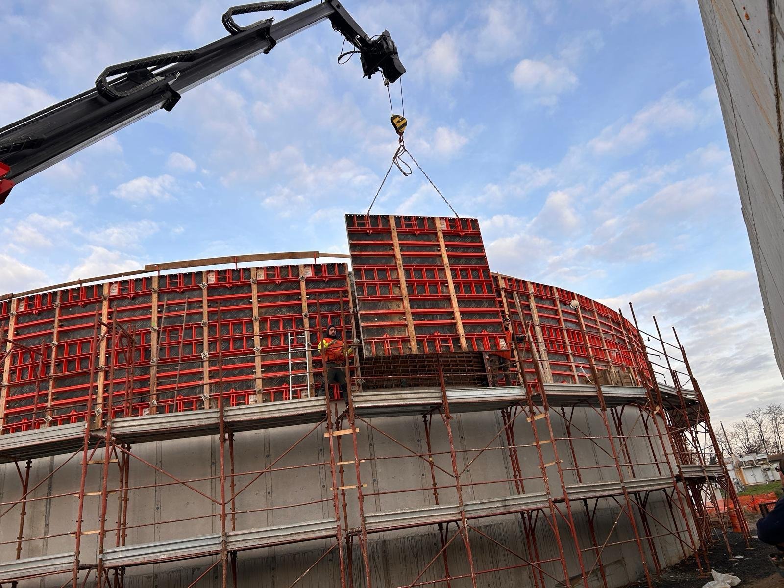 Construction workers building a curved concrete structure with red formwork and scaffolding, while a crane lifts a large red panel to the top of the structure under a cloudy sky.