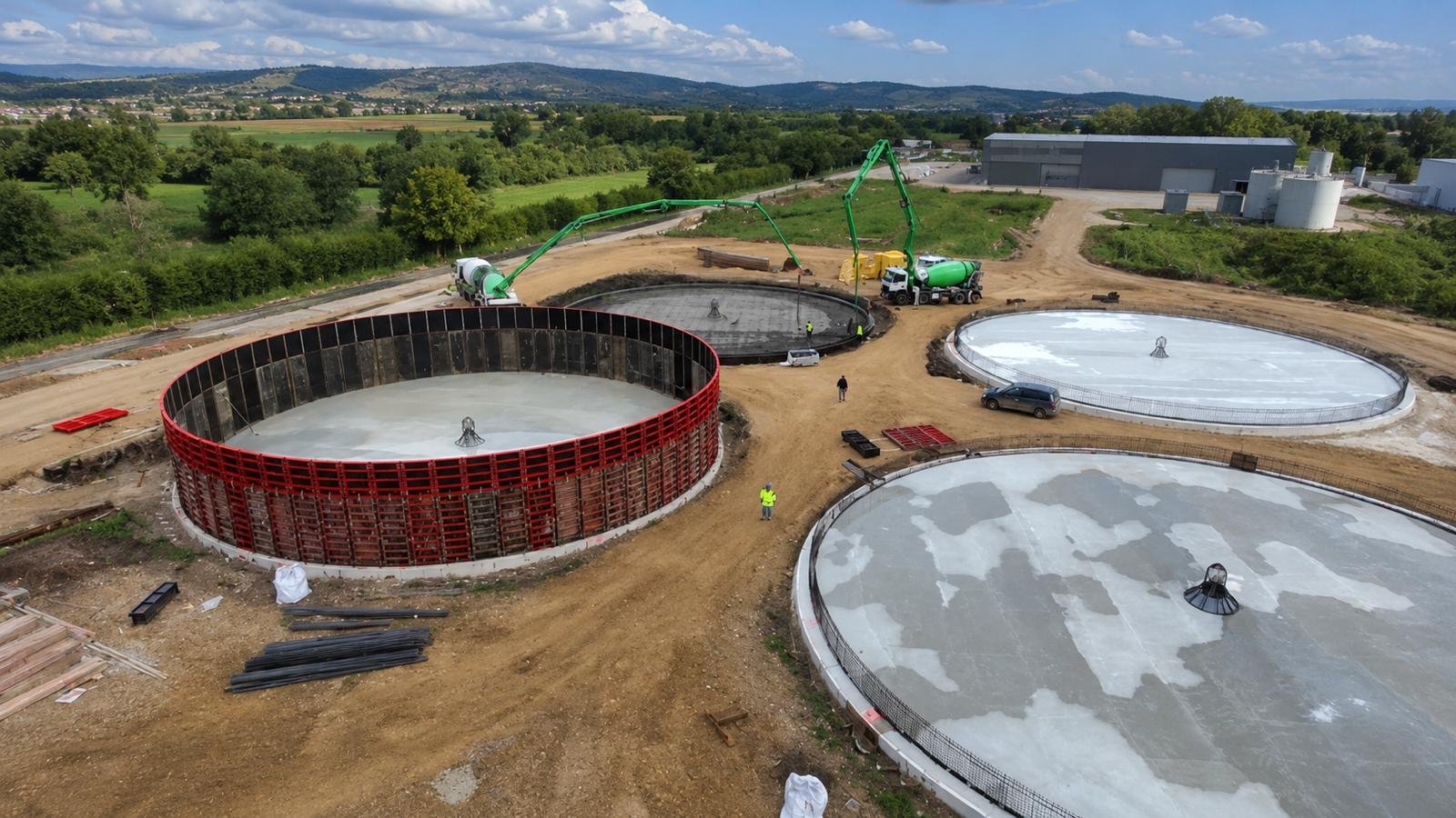 Construction site with large circular concrete tanks being built, with workers and construction equipment, in a rural setting with hills and green trees in the background.