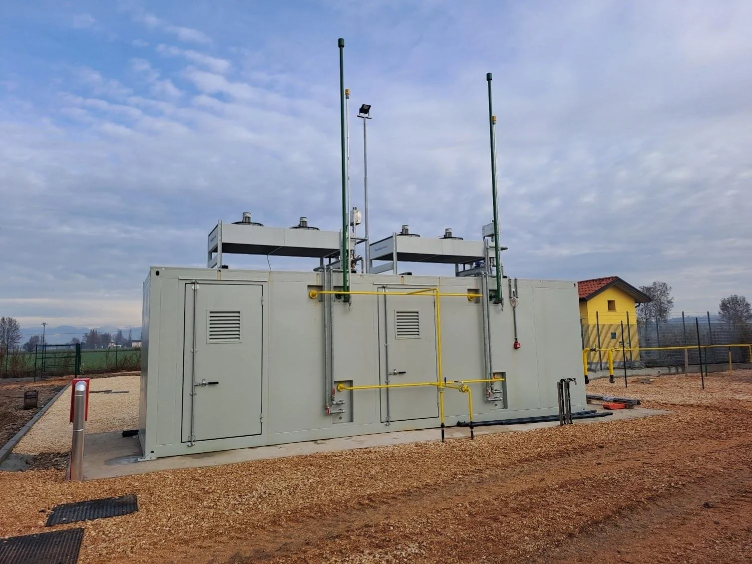 A large industrial electrical transformer with gray metal panels and yellow piping, located outdoors on a concrete pad, with a small yellow building and a chain-link fence in the background under a partly cloudy sky.