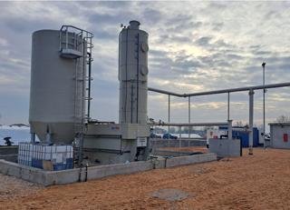 Industrial facilities with large cylindrical tanks, piping, and equipment on a dirt ground under a cloudy sky.