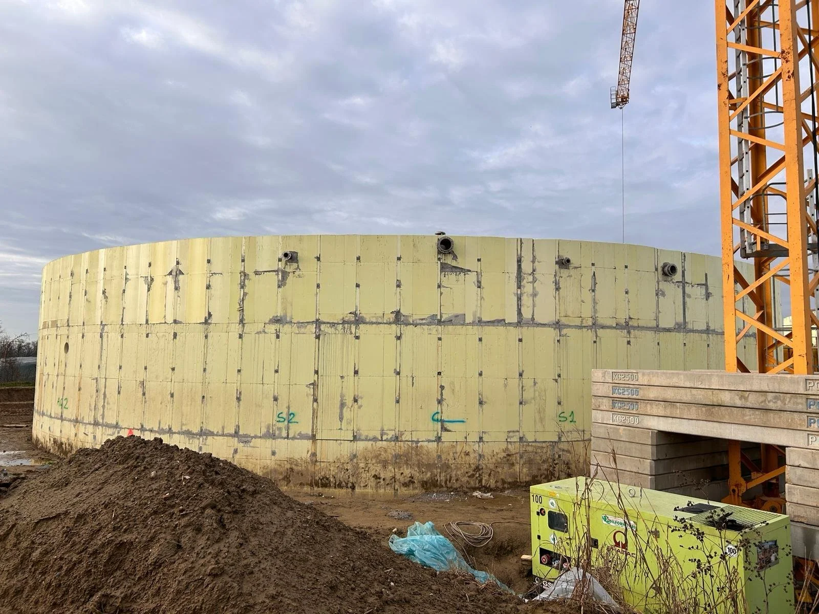 Construction site with a partially built circular structure, yellow insulation panels, a tall orange crane, and construction equipment, under a cloudy sky.