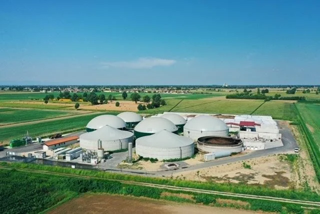 Aerial view of a cluster of large white industrial storage tanks in a rural area, surrounded by green fields and farmland under a clear blue sky.