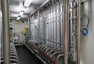 Industrial piping system with multiple vertical pipes and valves in a gray utility room.