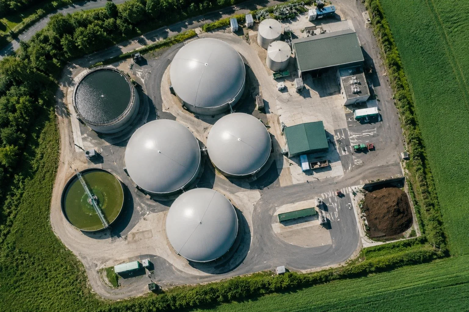 Aerial view of a biogas plant with multiple large, round storage tanks, some covered with metal domes, in a rural area surrounded by green fields and trees. The plant has a parking lot with some vehicles and machinery.