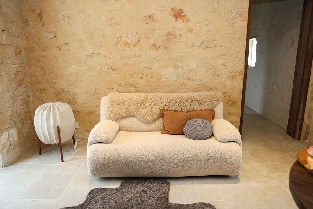 A beige two-seater sofa with three pillows, one brown, one gray, and one round, is placed against a textured stone wall in a room with beige tiled flooring and a small side table with a white lampshade to the left.