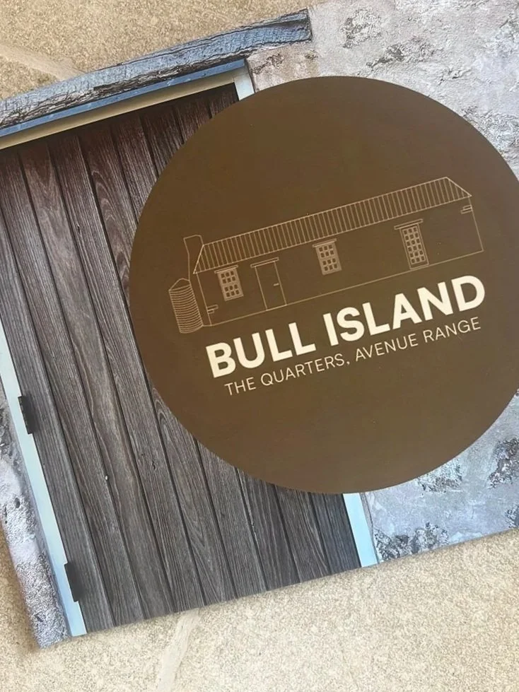 A circular sign with a line drawing of a building and the text 'Bull Island, The Quarters, Avenue Range' overlaying a wooden door and stone wall.