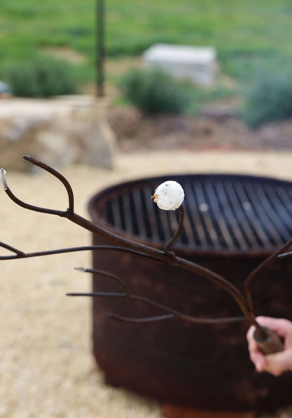 Close-up of a rusty metal fire poker with a white marshmallow skewered on its tip, held over a round grill in an outdoor setting.