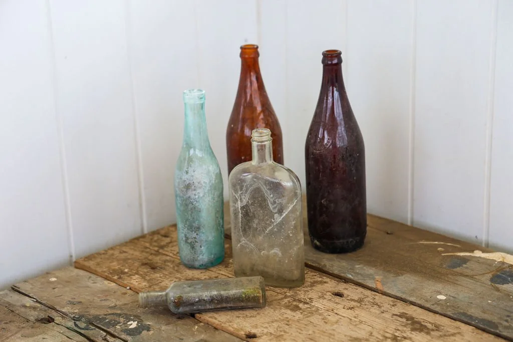 Five old glass bottles in different colors on a wooden surface against a white paneled wall.