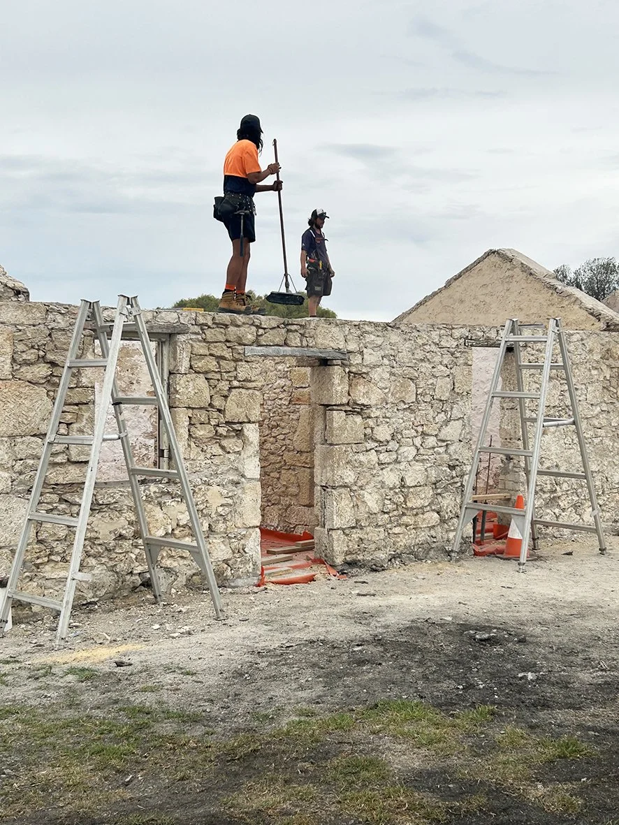 Two workers working on the top of a stone structure, with ladders and construction cones around, during daytime with cloudy sky.