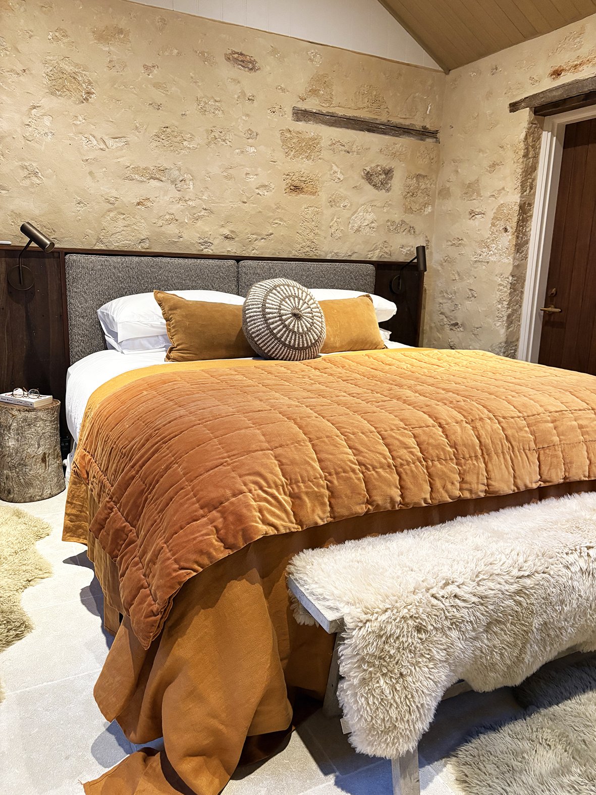 A cozy bedroom with a large bed covered in an orange quilt and pillows, against a textured stone wall, with a sheepskin-covered bench at the foot of the bed and a rustic wooden accent.
