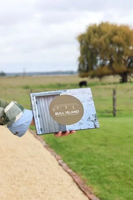 Person holding a cardboard box with a circular label that reads 'BULL ISLAND' outdoors on a farm with a dirt path, green grass, trees, and a cloudy sky in the background.