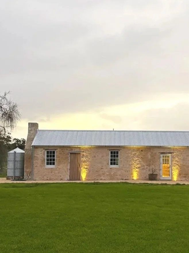 A stone building with a corrugated metal roof, lit with yellow lights at night, surrounded by a green lawn under a cloudy sky.