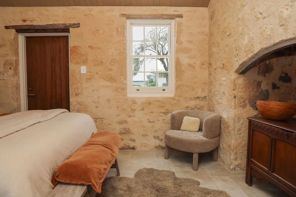 A cozy rustic bedroom with beige stone walls, a window showing a tree outside, a wooden door, a beige bed with an orange pillow, a small beige armchair with a beige cushion, and a wooden dresser with a bowl on top.