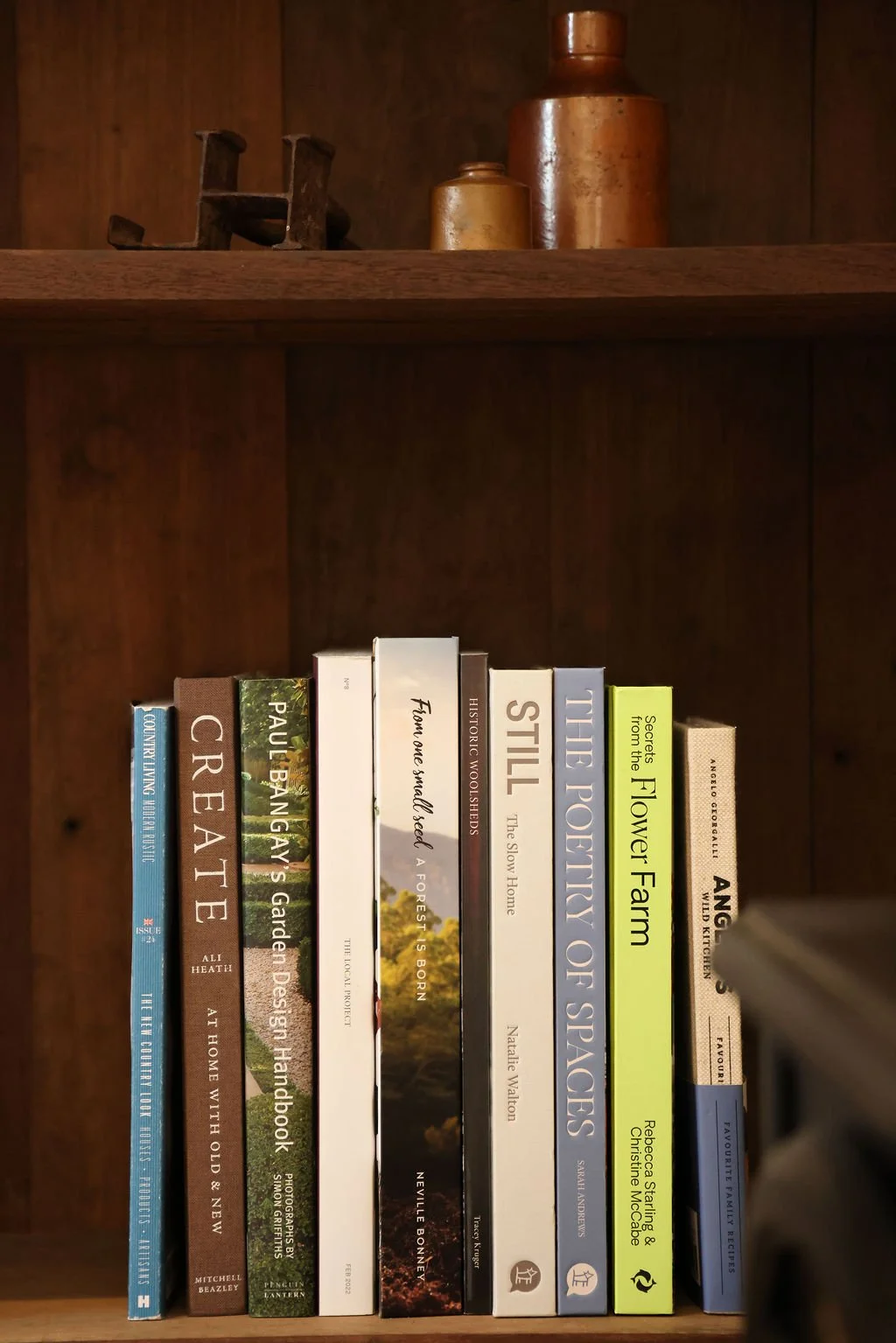 A wooden bookshelf with nine books and decorative items on top. The books include titles about gardening, history, and nature, with some titles visible such as "Create", "Paul Bangay's Garden Design Handbook", "Still", "The Poetry of Spaces", and "The Flower Farm". The items on top include a copper bottle, a smaller cup, and an old rusty printing press.