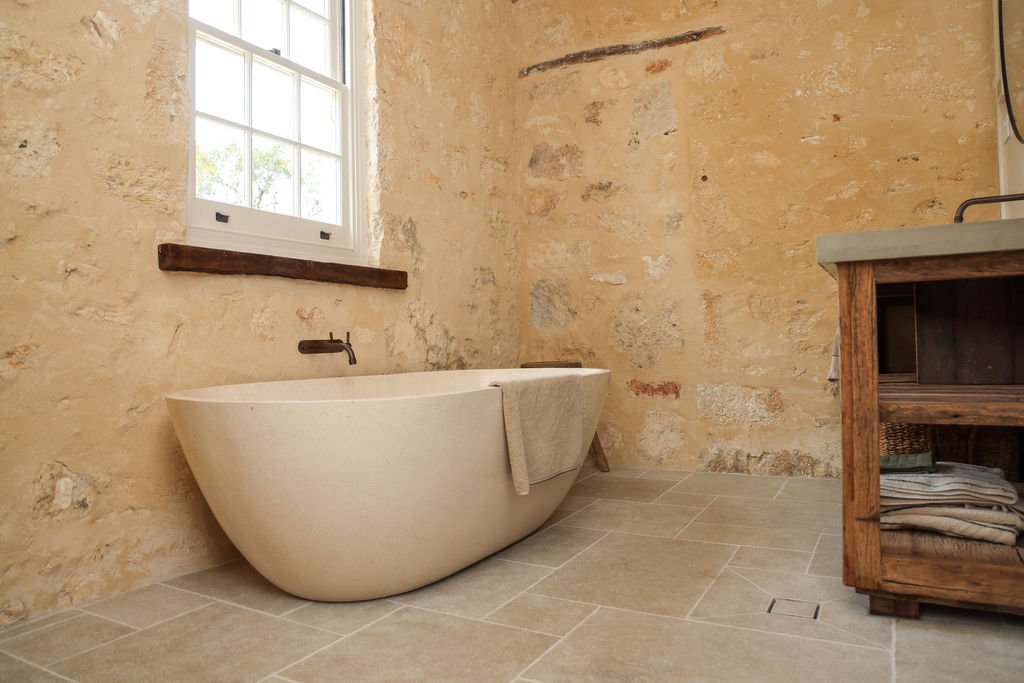 A rustic bathroom features a freestanding white bathtub, a stone wall, a window with white trim, and a wooden shelf with folded towels.