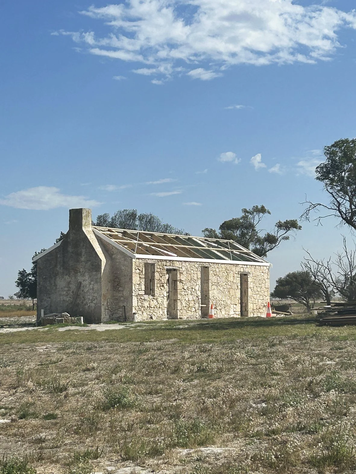Old stone building under renovation with a partially open roof, surrounded by open land and trees, with a blue sky and clouds overhead.