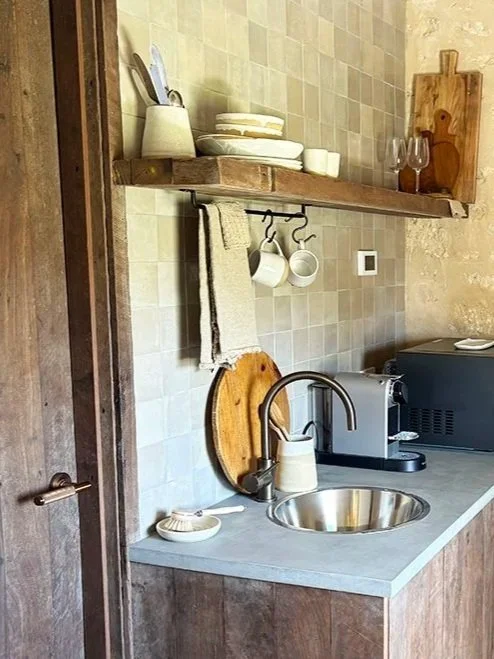 Small kitchen corner with a stainless steel sink, a faucet, a paper towel holder, and a dish sponge on the countertop. Open wooden shelf above with dishes, glasses, and kitchen utensils.