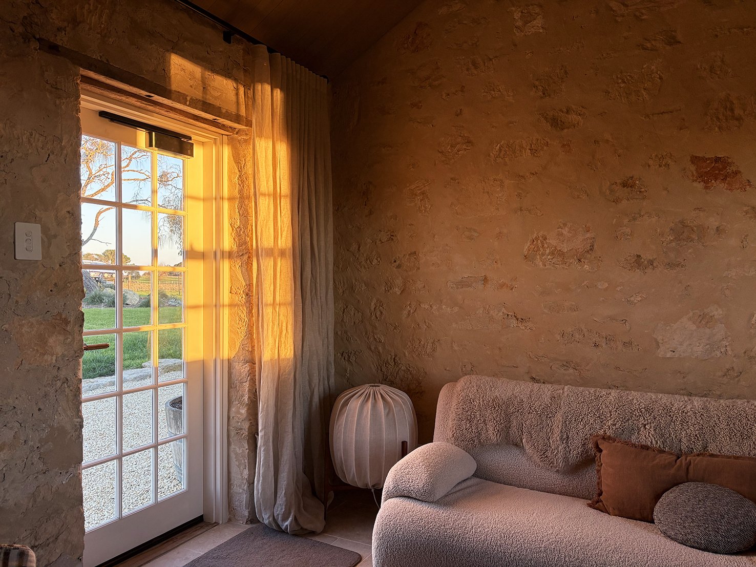 Sunlight casting a warm glow on a stone wall, a glass door with patterned panes leading outside, beige curtains, a rounded side lamp, and a cozy beige sofa with brown pillows inside a living room.