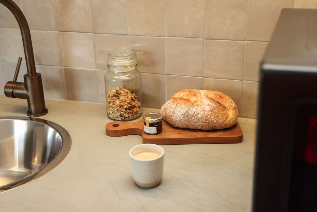 Fresh bread on a wooden cutting board, a jar of cereal, a small jar of jam, and a cup of coffee on a kitchen countertop with a metal sink and a tiled backsplash.