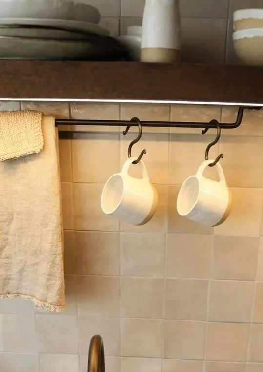 Two white coffee mugs hanging upside down on hooks beneath a kitchen shelf.