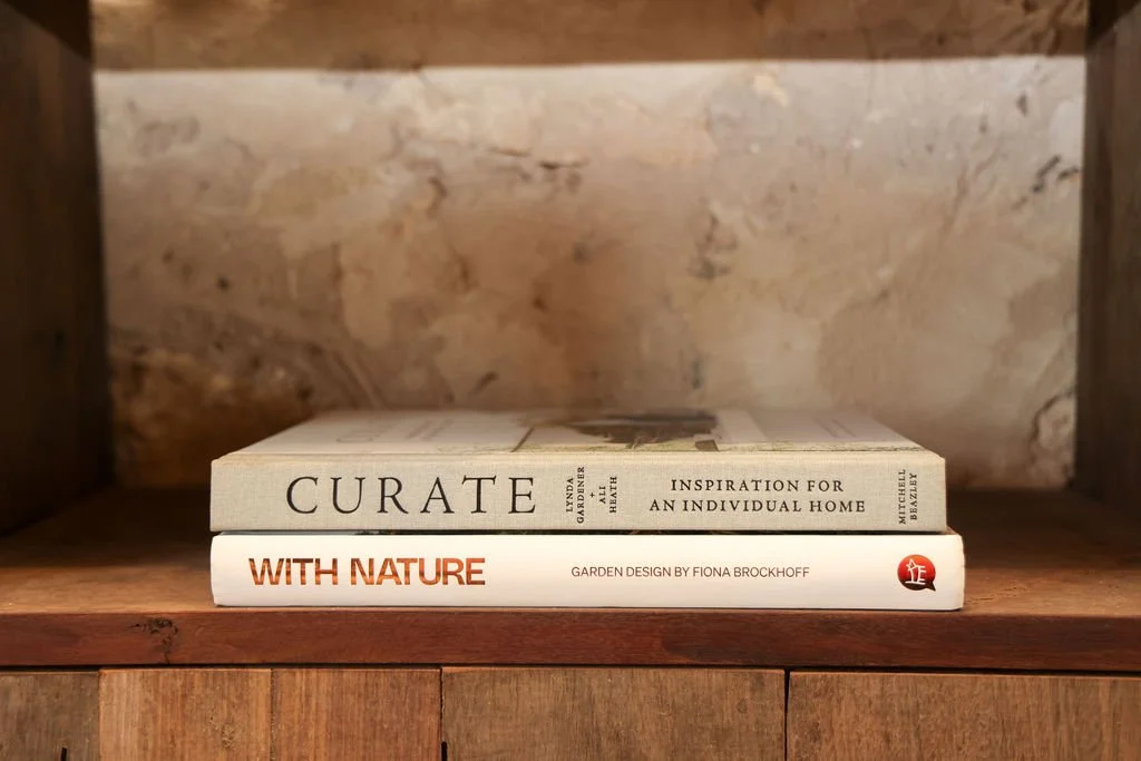 Two books stacked on a wooden shelf with a brick wall background. The top book is titled "Curate" and the bottom book is titled "With Nature" by Fiona Brockhoff.